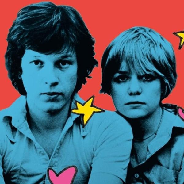 A portrait of Chris Frantz and Tina Weymouth sitting next to each other. Their bodies are colorized blue and the background is red. 