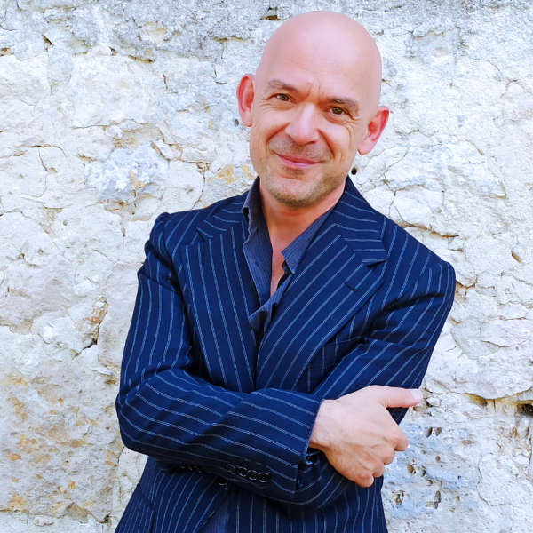 A man in a blue suit with his arms crossed smiling in front of a white wall
