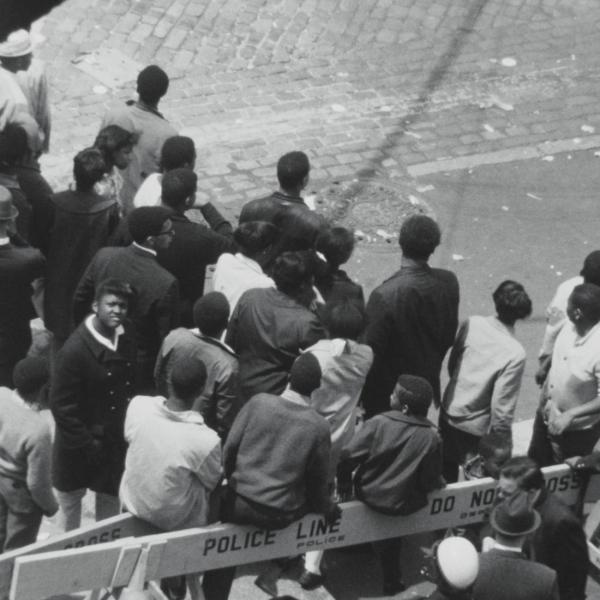 A black and white birdseye view of a group of people stand in the street. Police barricades delineate the space. 