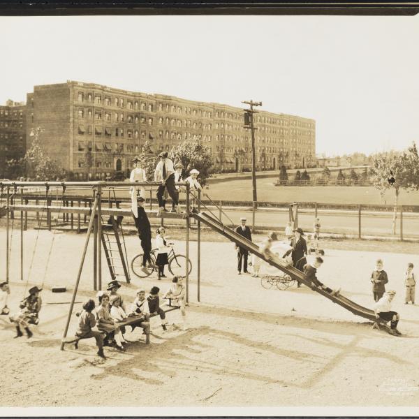 A black-and-white photograph from 1921 showing multiple children playing on a playground with swings, a slide, and a seesaw. Behind the playground is a street and apartment buildings.