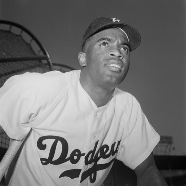 Baseball player Jackie Robinson wears his Brooklyn Dodgers uniform and hat, with a baseball stadium in the background