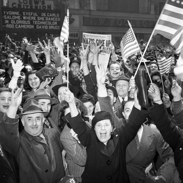 A crowd of men and women on Broadway in Manhattan hold American flags and throw their arms up celebrate the end of World War II in Europe.