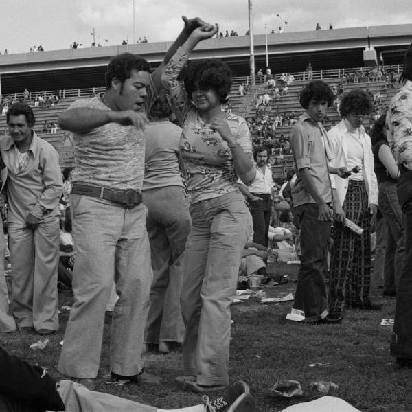 A casually dressed couple dances salsa with people in the background on Randall's Island in 1974.