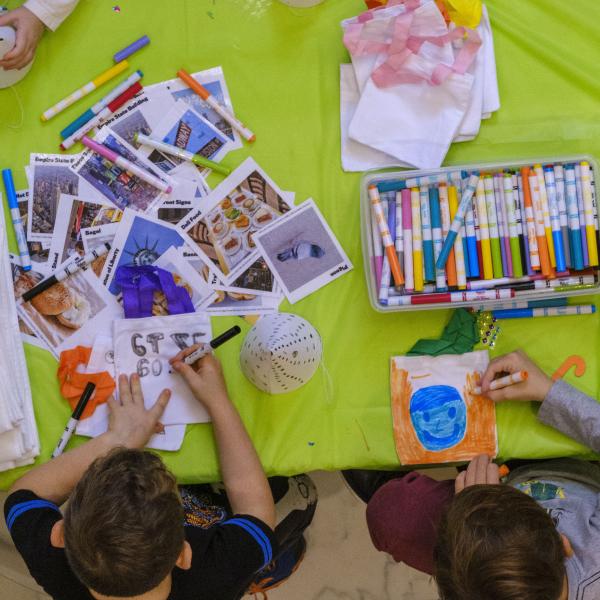 Foto aérea de duas crianças colorindo com marcadores e outros materiais de arte coloridos em uma mesa ao redor delas.