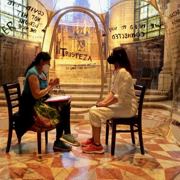 Dos mujeres sentadas y hablando dentro de una burbuja de plástico con mensajes escritos en las paredes.