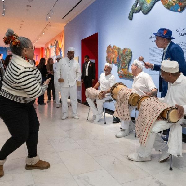 Três músicos vestidos de branco sentados em cadeiras tocando bateria latina. Mulheres dançando na frente de músicos. Artista Manny Vega em terno azul com instrumento shaker.