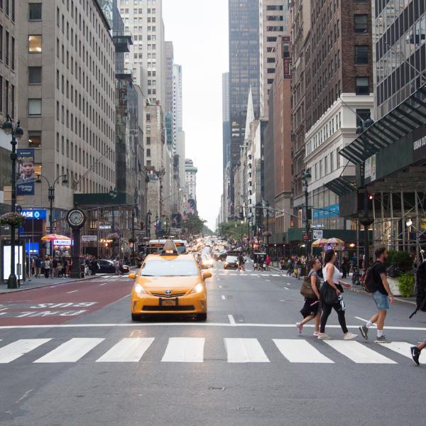 An intersection in NYC with a yellow taxi stopped at a light, and five pedestrians crossing the street. 