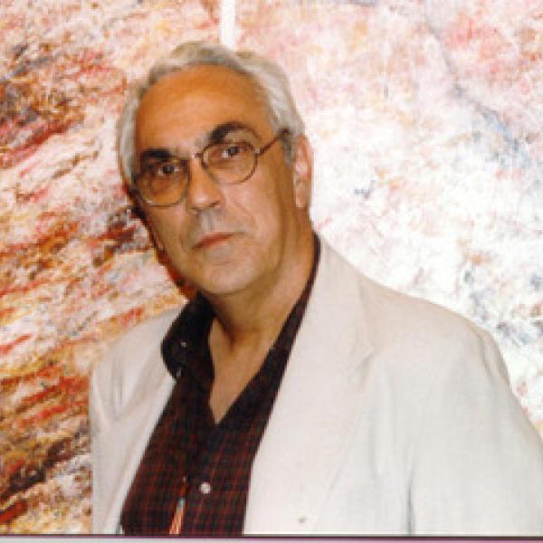 Color photograph of Mario César Romero wearing a white suit jacket and burgundy button down shirt against a brown-marbled wall