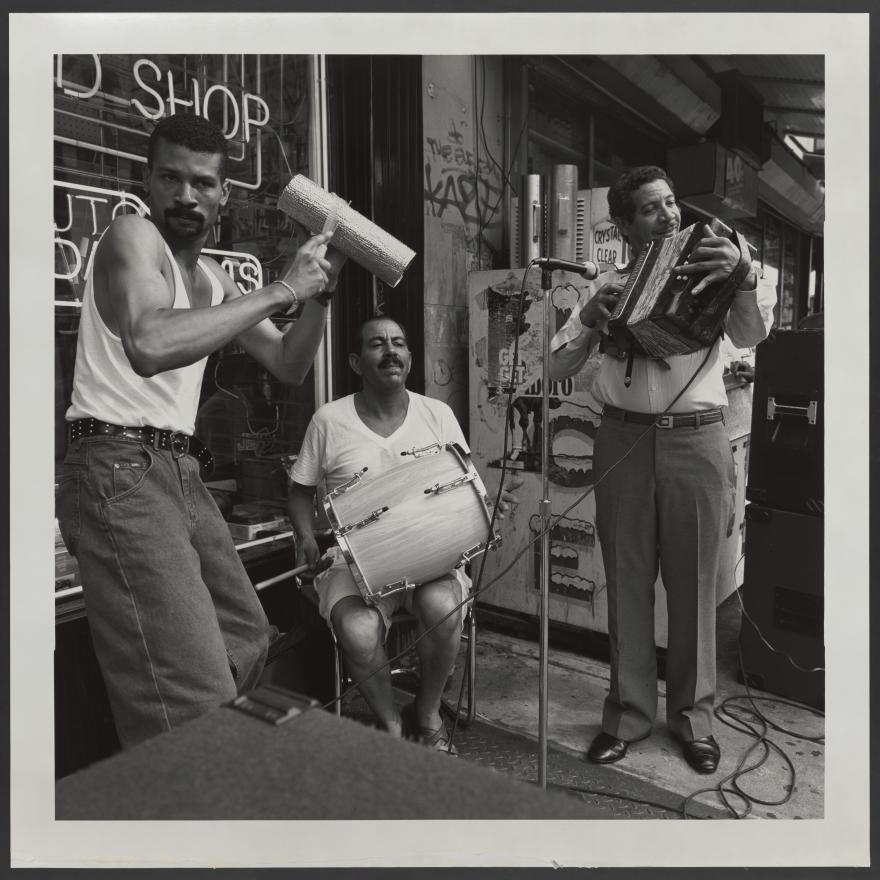 Black and white photograph of three men playing musical instruments outsde of a shop. One man is seated with a drum on his lap while the other two are standing, with one strumming a guira and another smiling as he plays his instrument.