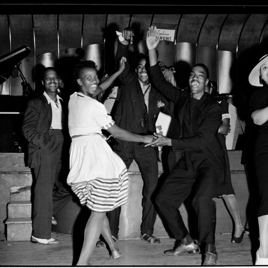 A lively group of Black men and women dance the Jitterburg and smile joyfully at nightclub in Harlem, 1939. A woman in a striped dress twirls with a partner at center, while others clap, cheer, and dance nearby. A jazz band is visible in the background, with a pianist and a saxophonist playing on stage. The atmosphere is energetic and celebratory. 