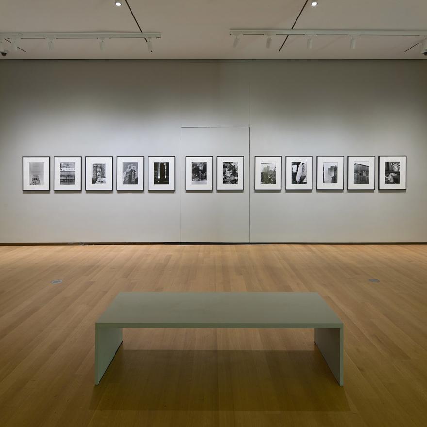 Installation of Robert Rauschenberg’s New York with a bench in front of a wall of photographs