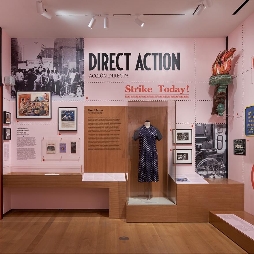 A gallery with pink walls and wooden cases showing photographs, sculptures, paintings, and clothing