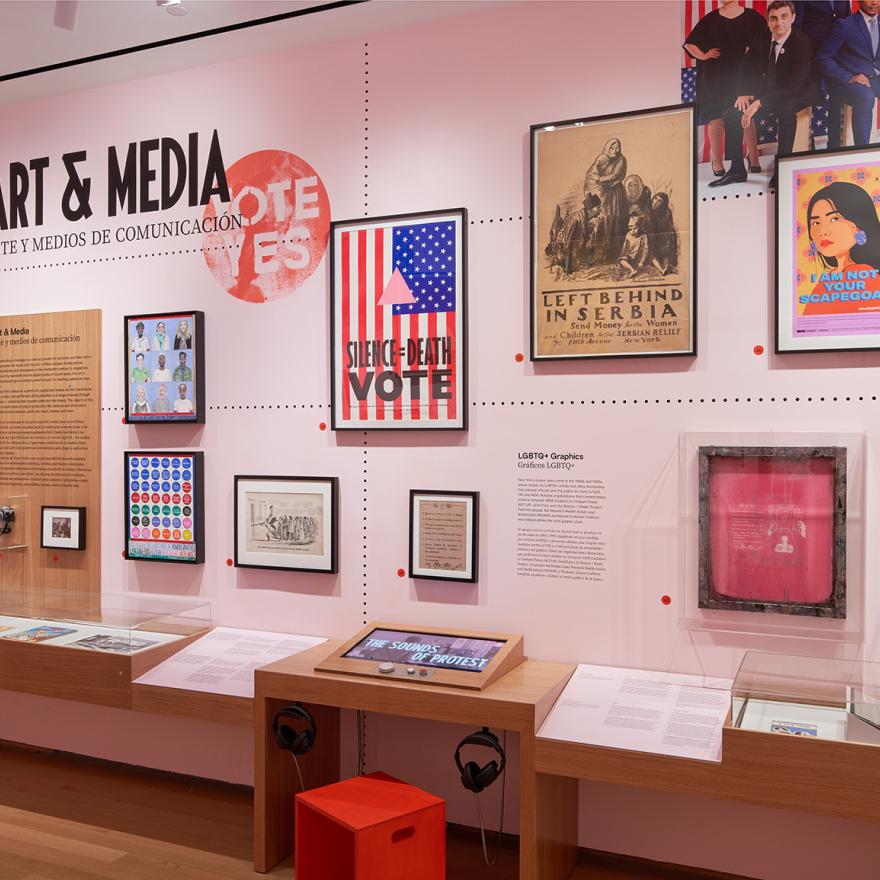 A gallery with pink walls and wooden cases showing photographs, sculptures, paintings, and interactive displays