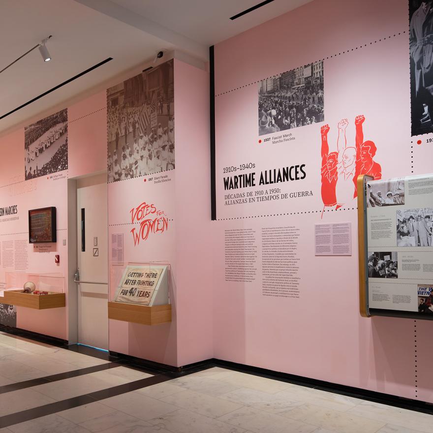 A gallery with pink walls and wooden cases showing photographs, sculptures, paintings, and interactive displays