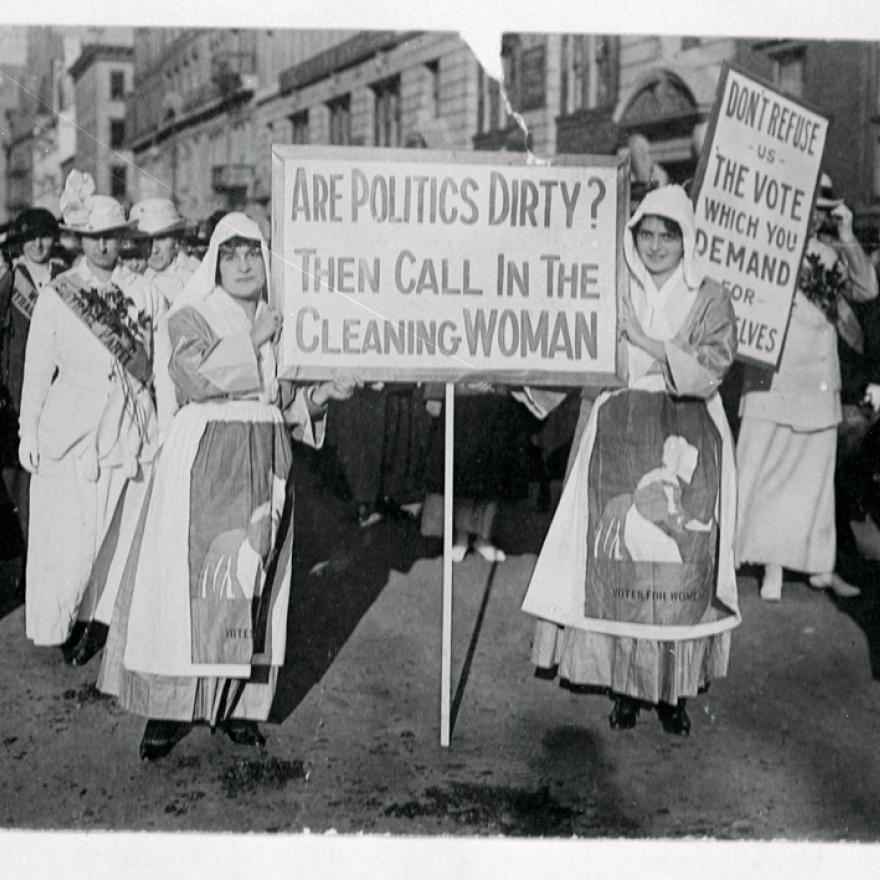 During a suffrage rally two women face the camera with a sign that says “Are politics dirty? Then call in the cleaning woman”