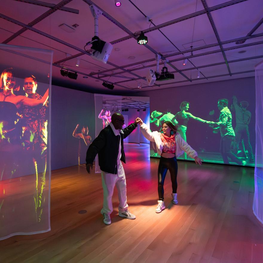 Two people dance together in a colorful, immersive art installation. The room features wooden floors and sheer fabric panels hanging from the ceiling, onto which vibrant, life-size video projections of dancers are displayed. The walls also show large-scale projections of people dancing, creating a lively, energetic atmosphere. Overhead lighting adds to the vivid, multicolored ambiance of the space.