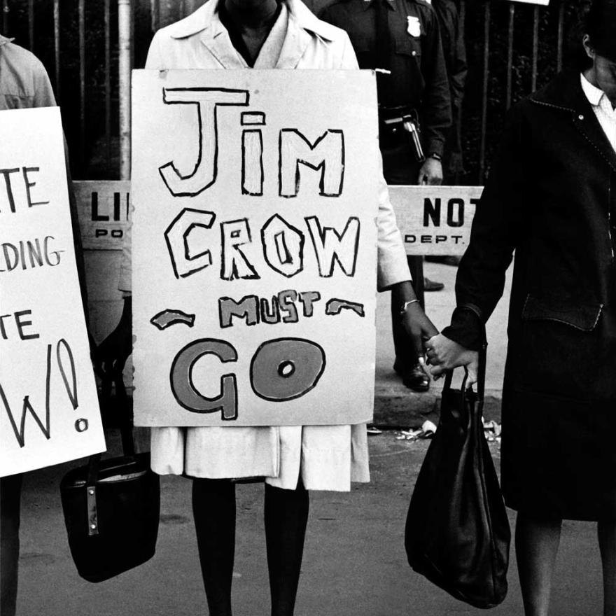 A woman stands in a line of protesters holding hands, with a sign around her that says “Jim Crow must go”