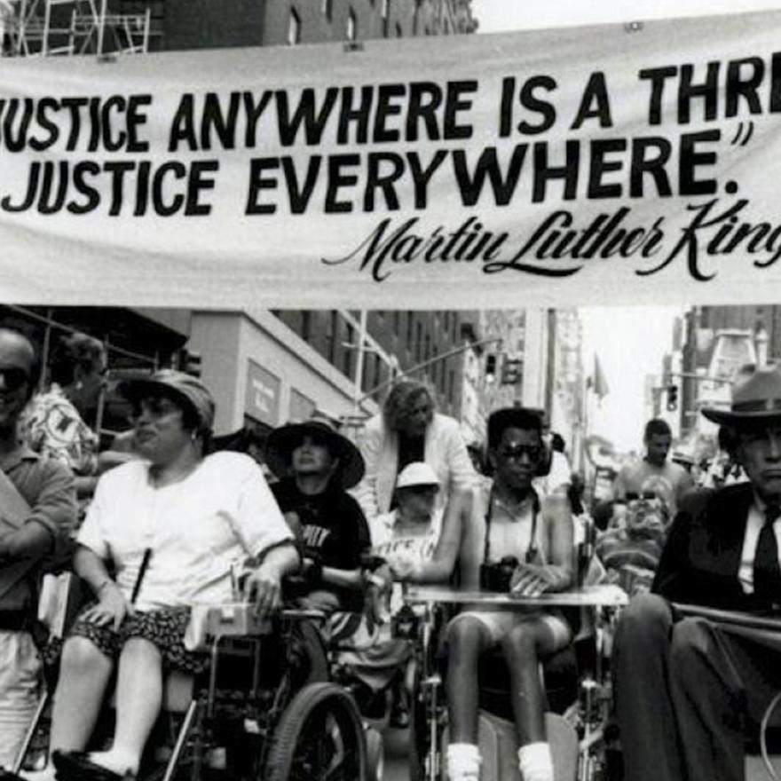 A crowd of people with disabilities and individuals in wheelchairs gather under a banner that reads "Injustice Anywhere is a Threat to Justice Everywhere" Martin Luther King Jr.