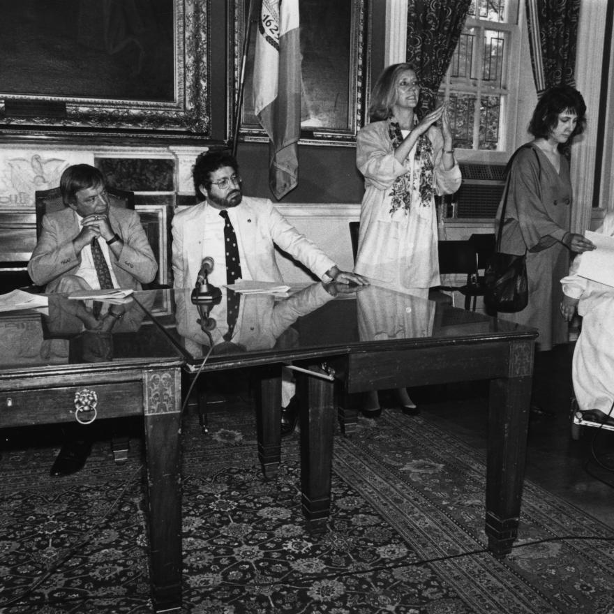 En una habitación profusamente decorada, tres hombres con traje se sientan en mesas a la izquierda con micrófonos. A la derecha, una mujer se pone de pie, firmando, mientras que más a la derecha, otra mujer sostiene un papel/discurso para una tercera mujer en silla de ruedas, que habla en un pie de micrófono.