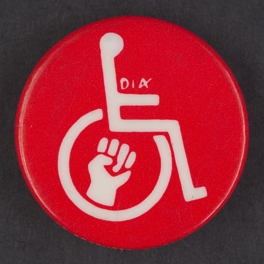 Circular metal button. Red with a white symbol of an outline of a person in a wheelchair. Also in white is a symbol of a fist inside the wheel and the letters "D.A." in small type above the arm.
