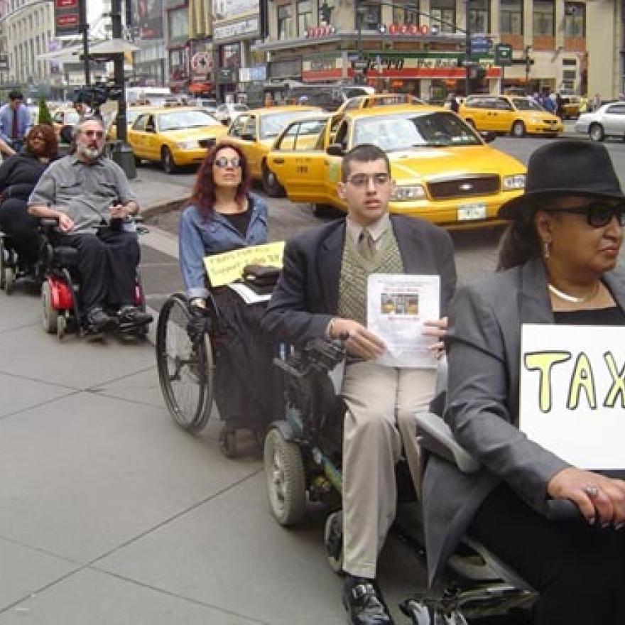 People in wheelchairs are lined up at the edge of a street with signs that say "TAXI." In the street next to them is a row of taxi cabs.
