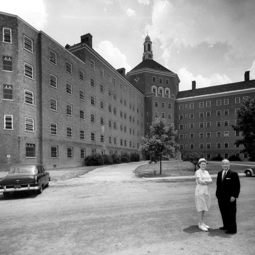 Una enfermera con un vestido y un sombrero blancos se encuentra junto a un hombre con un traje oscuro en el extremo derecho del primer plano de una fotografía en blanco y negro. Detrás de ellos, ocupando el fondo de la imagen, se encuentra un gran edificio de ladrillo.