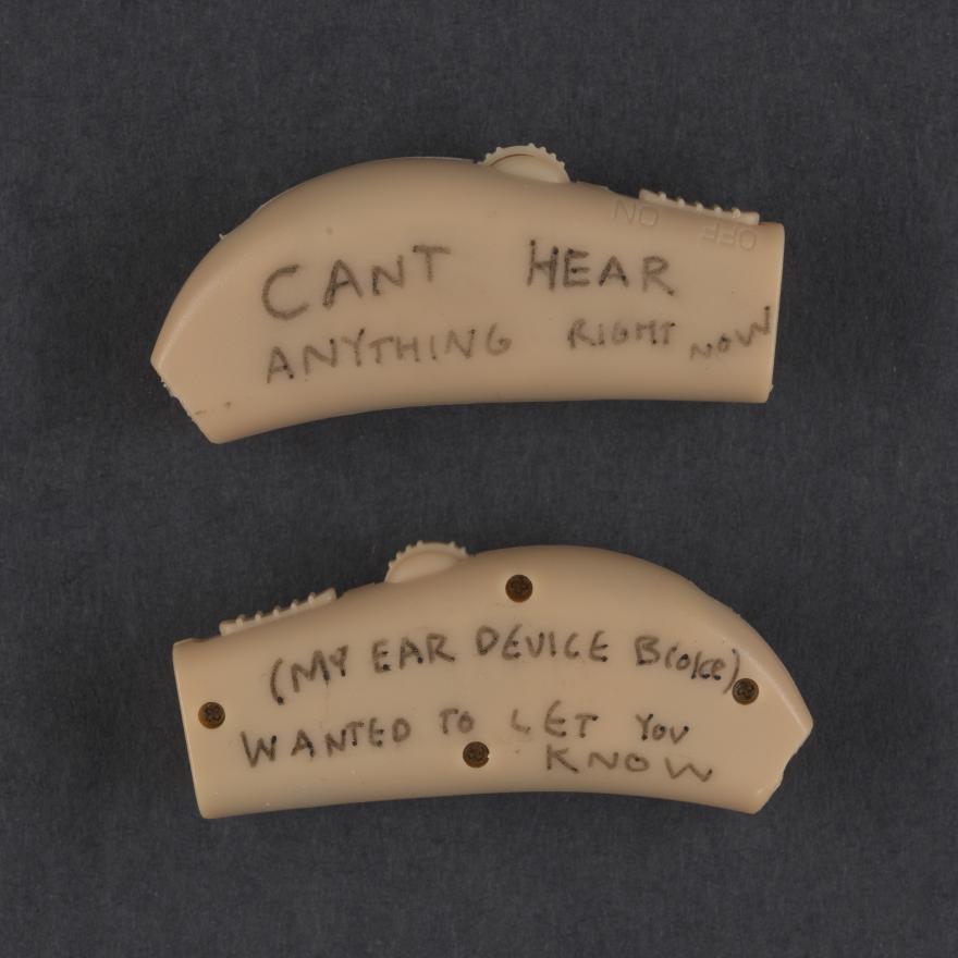 Image of a pair of small beige hearing aids with writing on them. Top reads: "CANT HEAR anything right now" bottom reads: "(MY EAR DEVICE BROKE) wanted to let you know