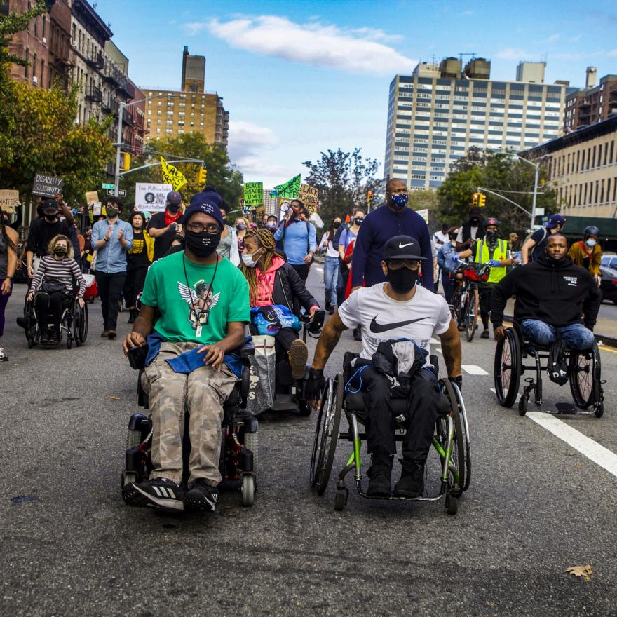 Two men in wheelchairs wearing masks lead a crowd of people, some in wheelchairs or with canes, some holding posters.