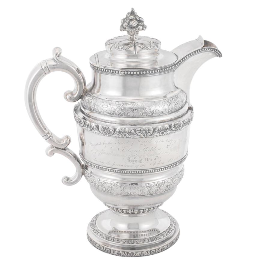 Silver pitcher with intricate details and writing in the middle