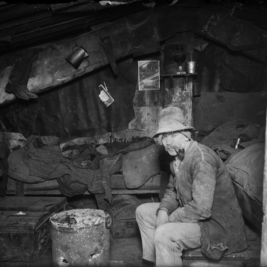 Man sits in a shack-looking house. Both the house and man are dirty, and look uncared for