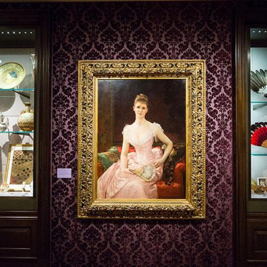 Two exhibition cases containing dishware and fans. In-between them is a painting of a seated woman in a pink dress