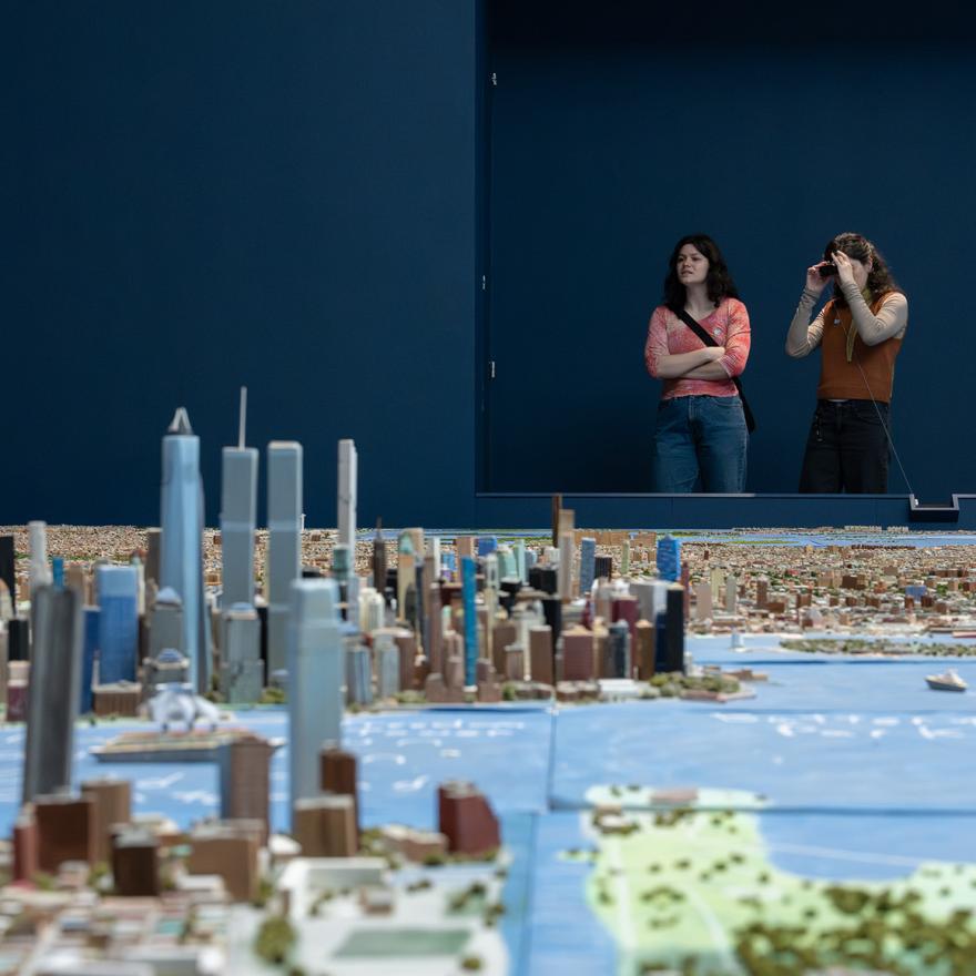 Two people looking at a handmade scale model of New York City through binoculars