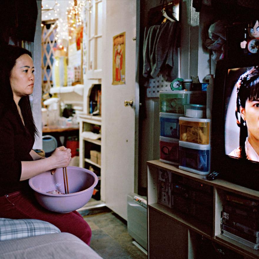 Uma mulher chinesa mexe comida em uma tigela enquanto assiste a uma novela chinesa. Ela está sentada em uma cama em um apartamento.