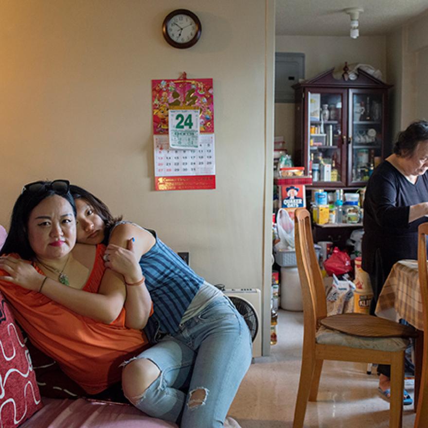 A woman sits on a couch, with her teenage daughter on her lap hugging her. An older woman stands in the background.