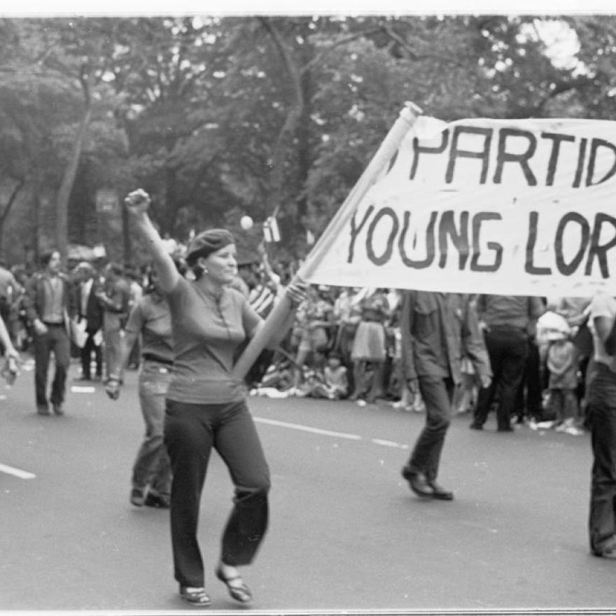 People march during a parade, while two hold a sign between them that says “_PARTIDO/YOUNG LORD”