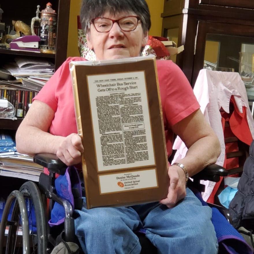 Una mujer con gafas en silla de ruedas sostiene un artículo de noticias enmarcado.