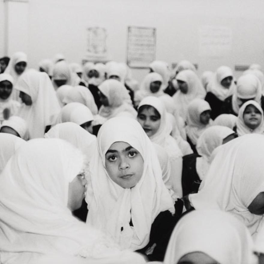 A group of girls wearing hijabs stand in a schoolyard, one girl in the middle stares into the camera