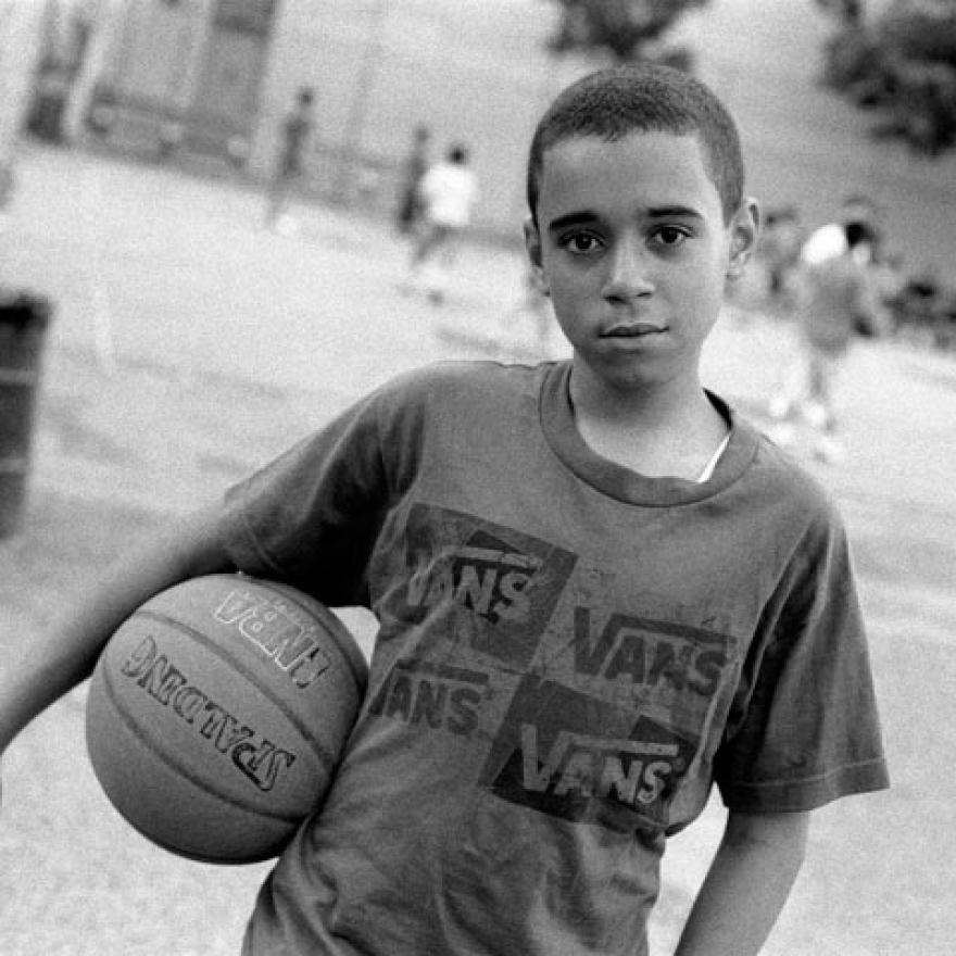 A boy stares into the camera while holding a basketball. Behind him, other children are playing basketball on a court