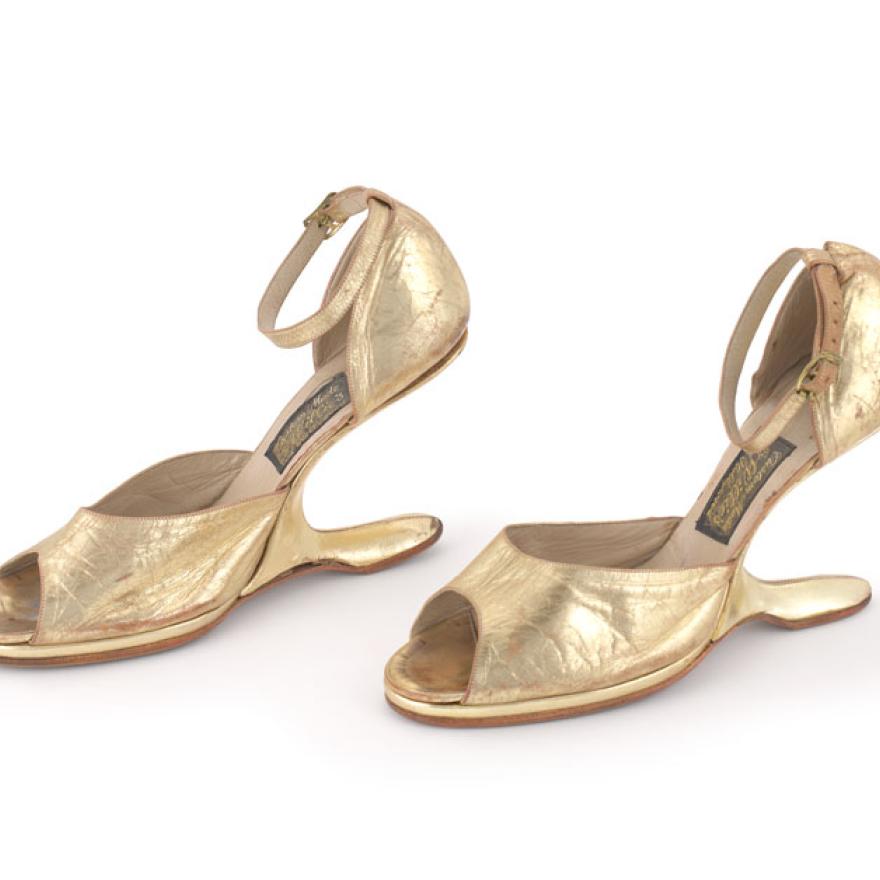 A pair of gold shoes, that are shaped like high heels. Rather than a heel, they have a piece that extends back from the base