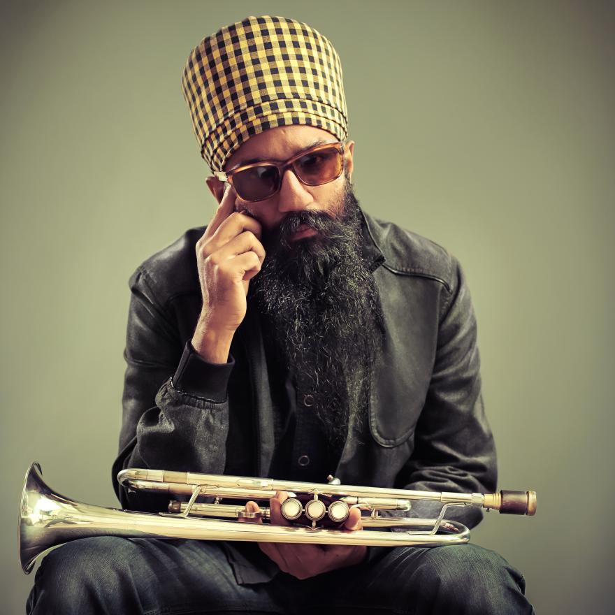 A man poses while wearing a checkered turban and holding a trumpet.  