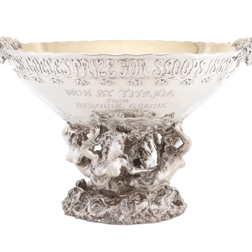 A large silver bowl held by statues of mermaids, with heads as handles and text on the bowl