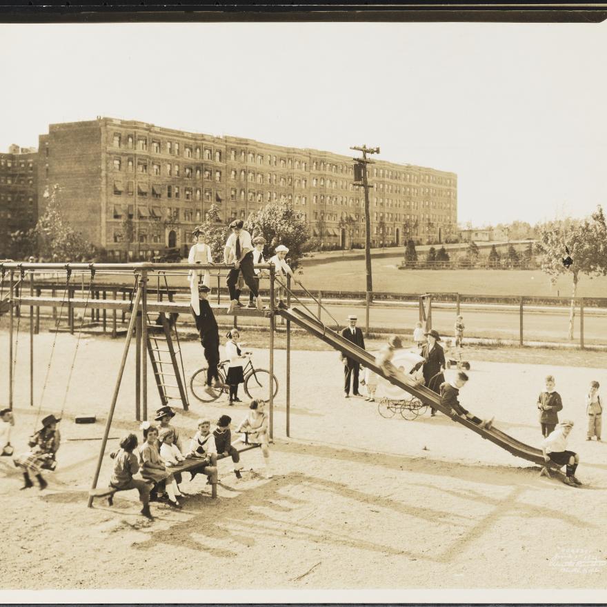 A black-and-white photograph from 1921 showing multiple children playing on a playground with swings, a slide, and a seesaw. Behind the playground is a street and apartment buildings.