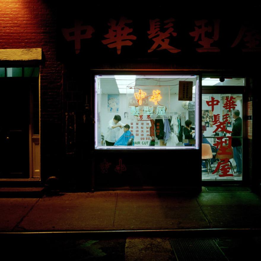 Taken from the street, a boy is seen getting his hair cut in a Chinese barbershop. The signs for the shop are in Chinese.