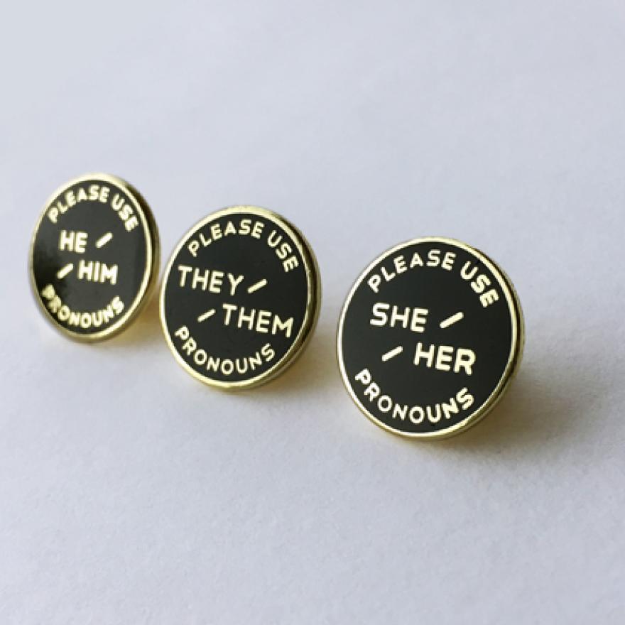 Three enameled pronoun pins. Black background with gold text. 