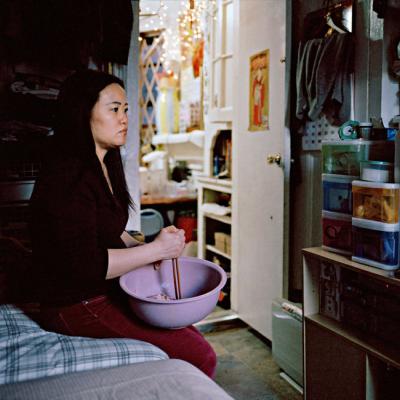 A Chinese woman stirs food in a bowl while watching a Chinese soap opera. She is sitting on a bed in an apartment.