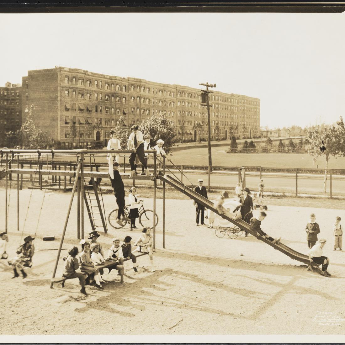 A black-and-white photograph from 1921 showing multiple children playing on a playground with swings, a slide, and a seesaw. Behind the playground is a street and apartment buildings.