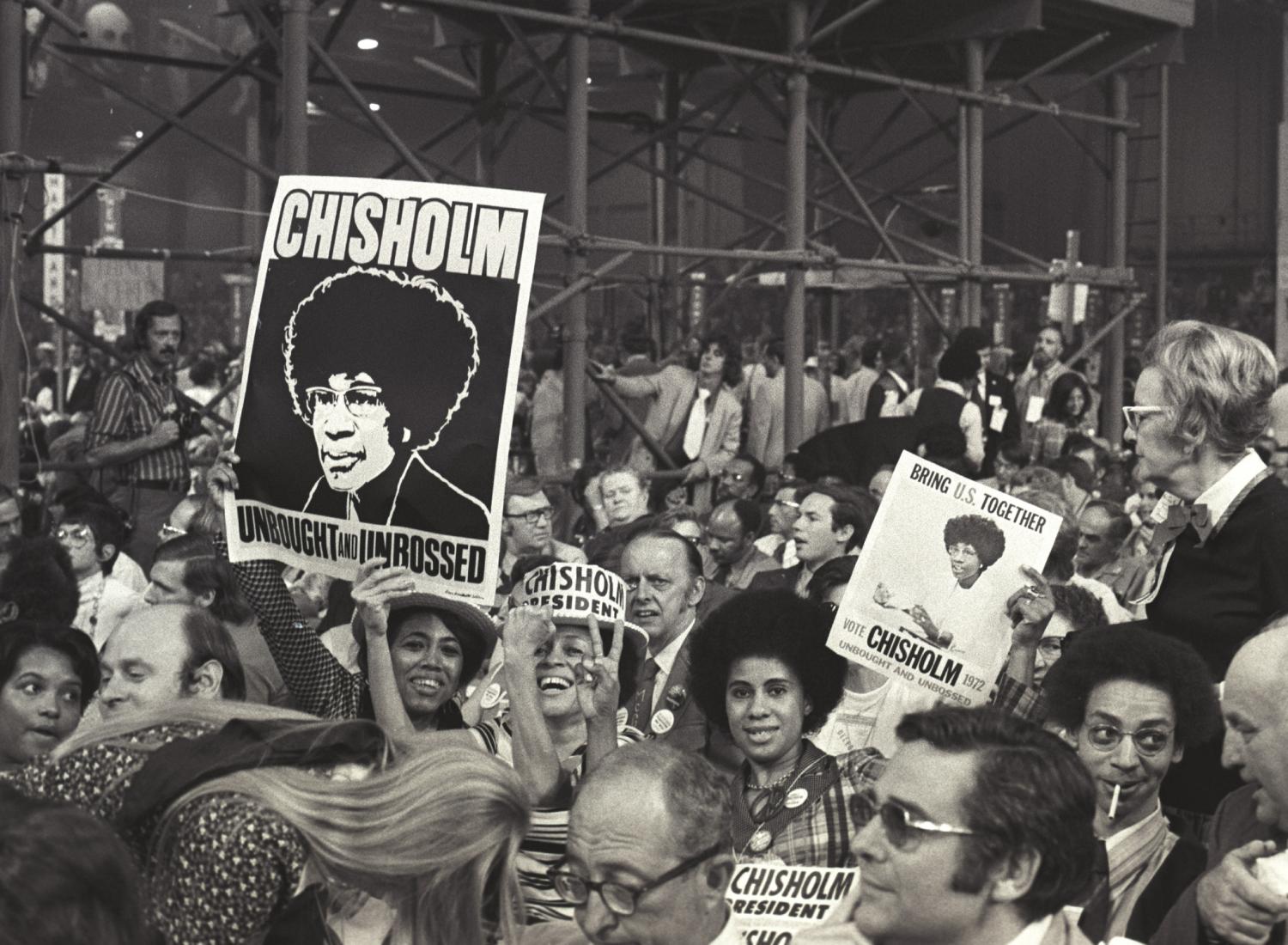Uma fotografia em preto e branco de uma multidão reunida em apoio à candidata Shirley Chisholm. Várias pessoas estão sorrindo e segurando cartazes apoiando sua campanha para presidente.