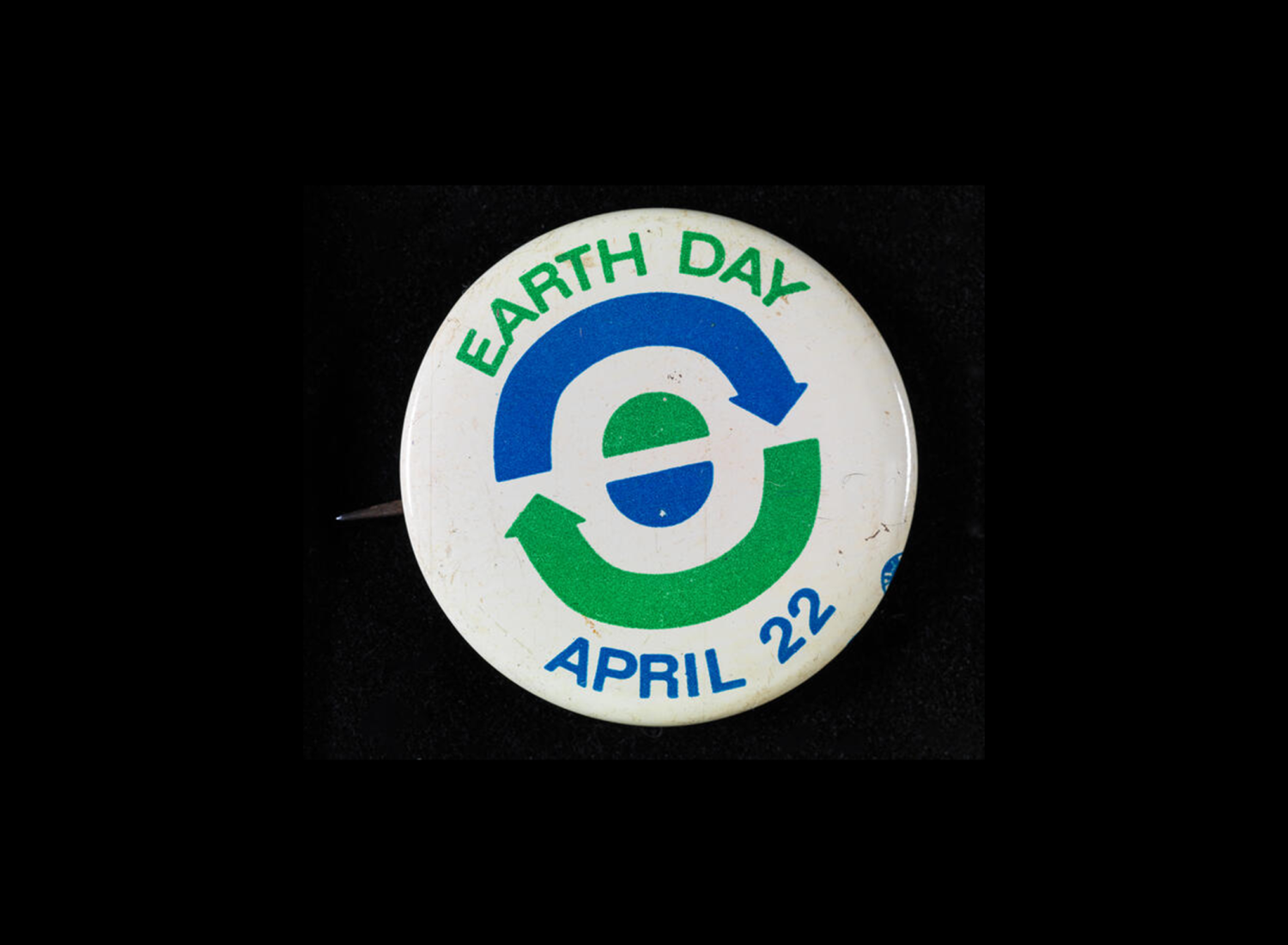 Um botão branco onde se lê Dia da Terra, 22 de abril, com setas azuis e verdes e semicírculos que simbolizam a reciclagem e a terra