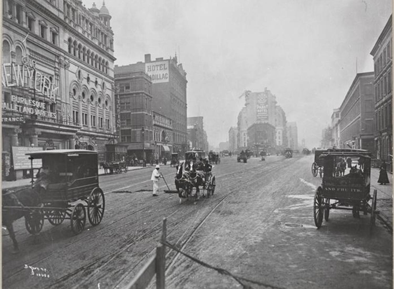 A black-and-white photograph showing the intersection of Broadway and 42nd Street, the present-day location of Times Square. The city street is surrounded by multistory buildings, and horse-drawn carriages drive in the street. In the center of the street is a New York City Sanitation worker dressed in white.
