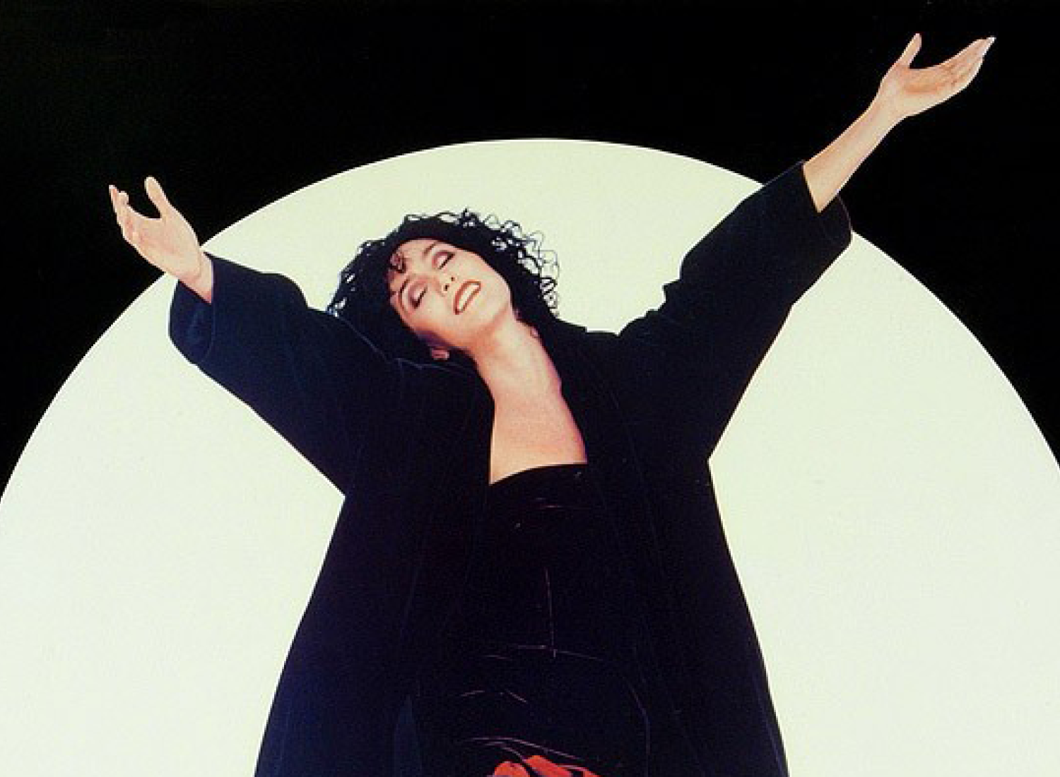 Cher ergue os braços no ar na frente de uma lua cheia.